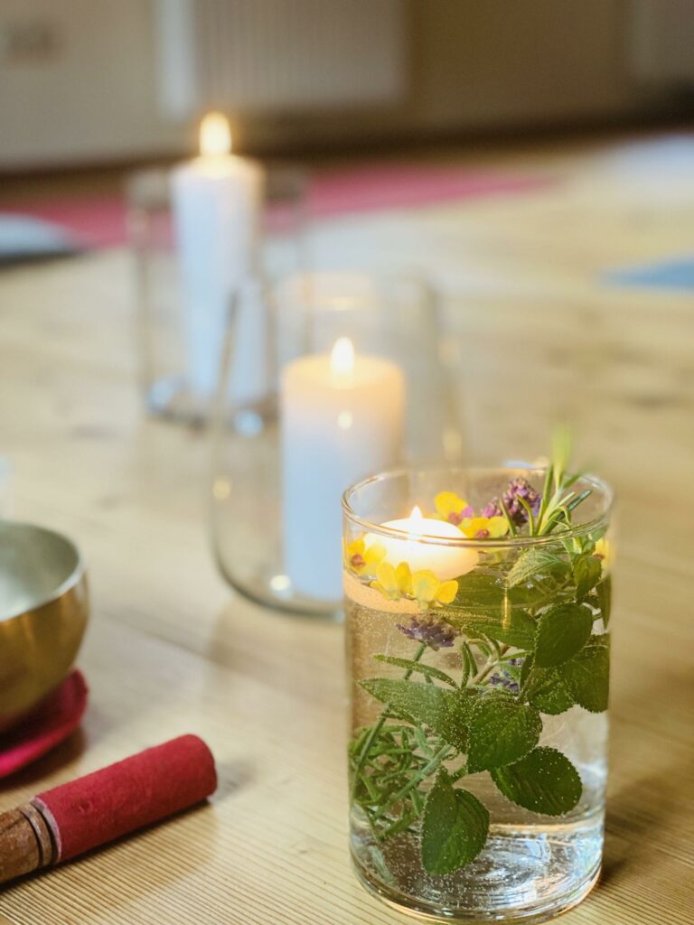 Kerzen und eine Glasschale mit Blumen und Kräutern neben einer Klangschale