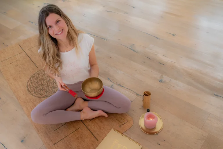 Yogalehrerin sitzt lächelnd mit einer Klangschale auf der Matte